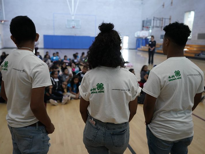 Three kids standing in front of a group of kids sitting on the floor of a gym.