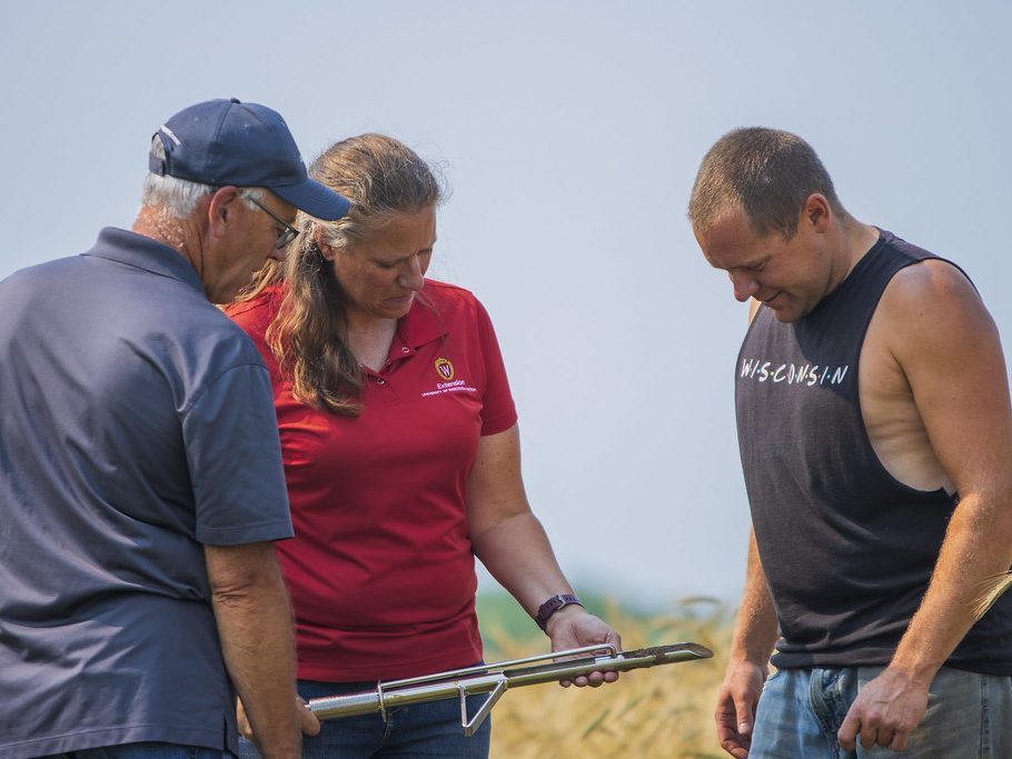 A woman holding an instrument that is put into the soil to grab a sample. Two men standing looking at the soil sample. 