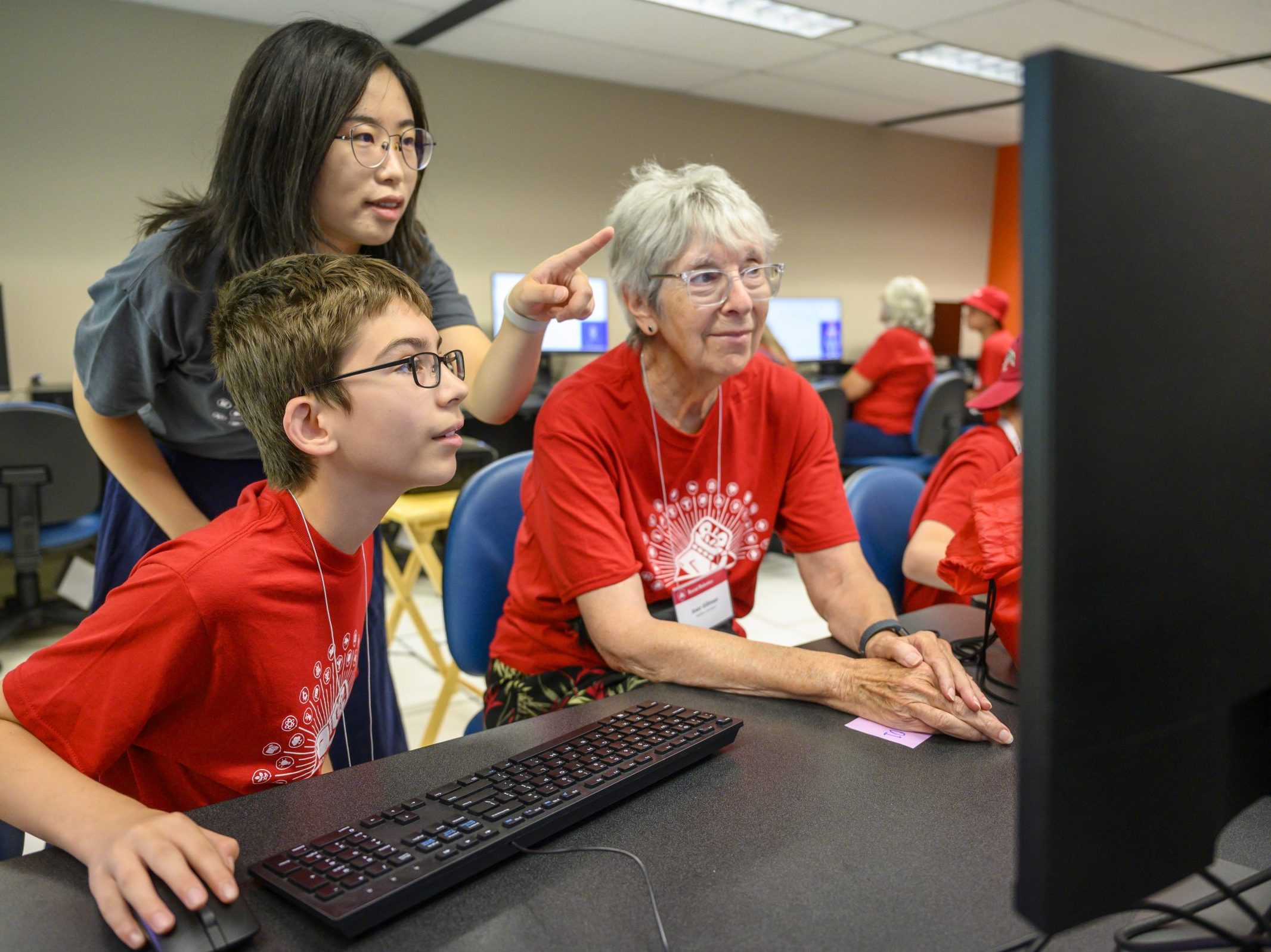 A boy looking at a computer screen with an older woman next to him. Another woman is standing over them both pointing at the screen.