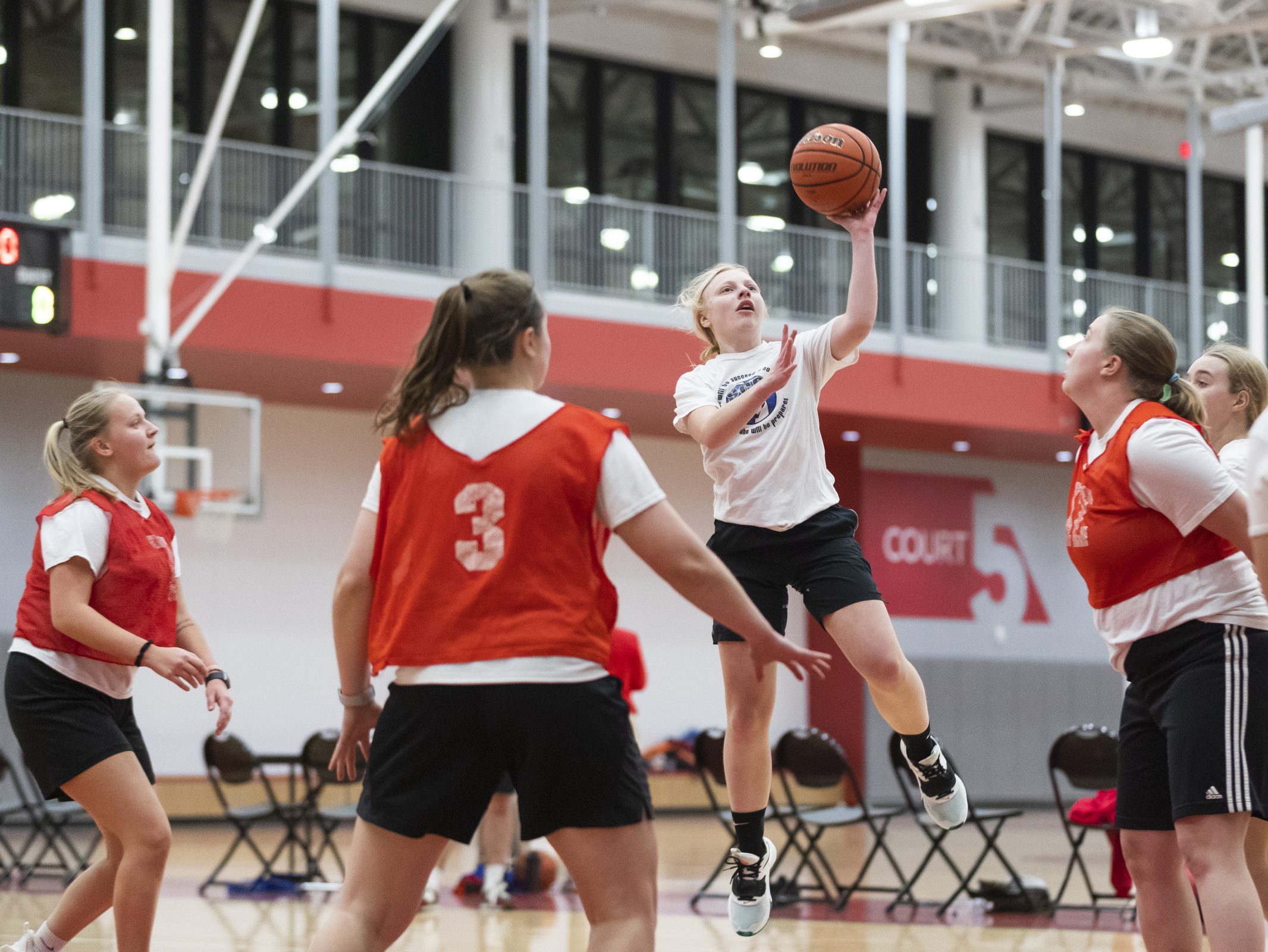 Girls in red jerseys surrounding a girl in white who is jumping up with a basketball in hand to make a basket