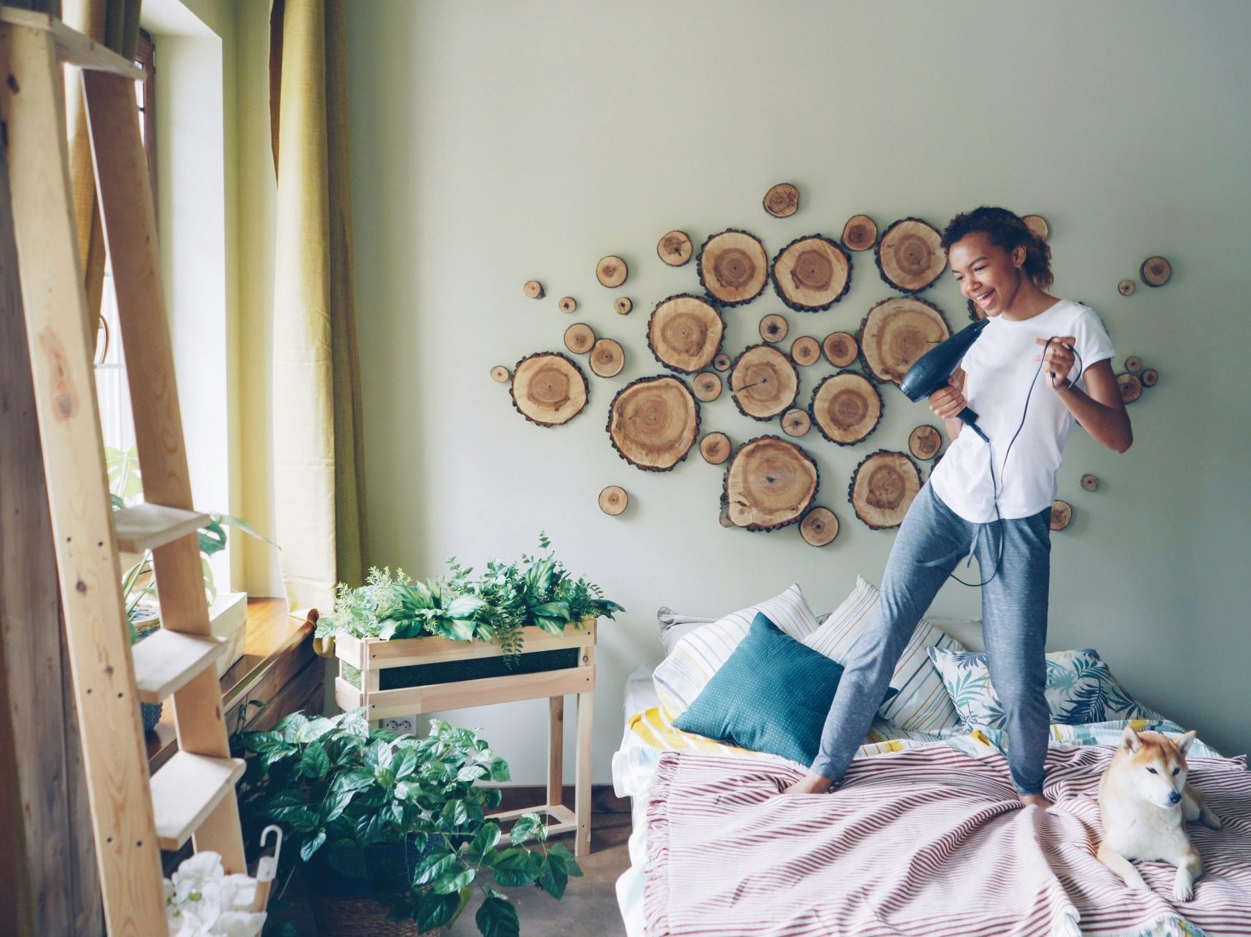 Girl on bed singing into a hair dryer. Room is decorated nicely with some plants, pink bed sheets, and wood slices creating a design on the wall.