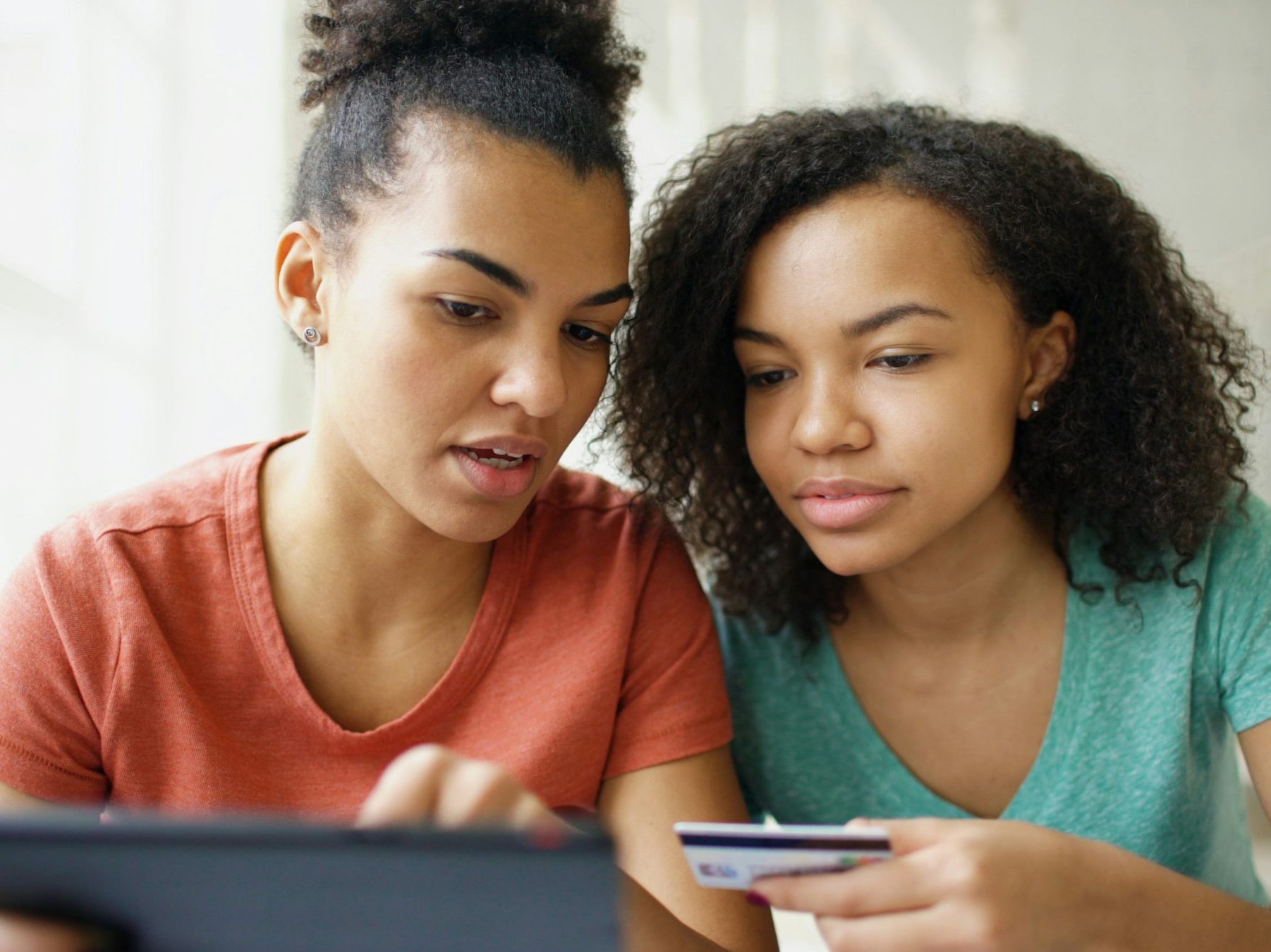 A mom and daughter at a tablet. Daughter has a credit card in hand putting in the numbers while mom watches.