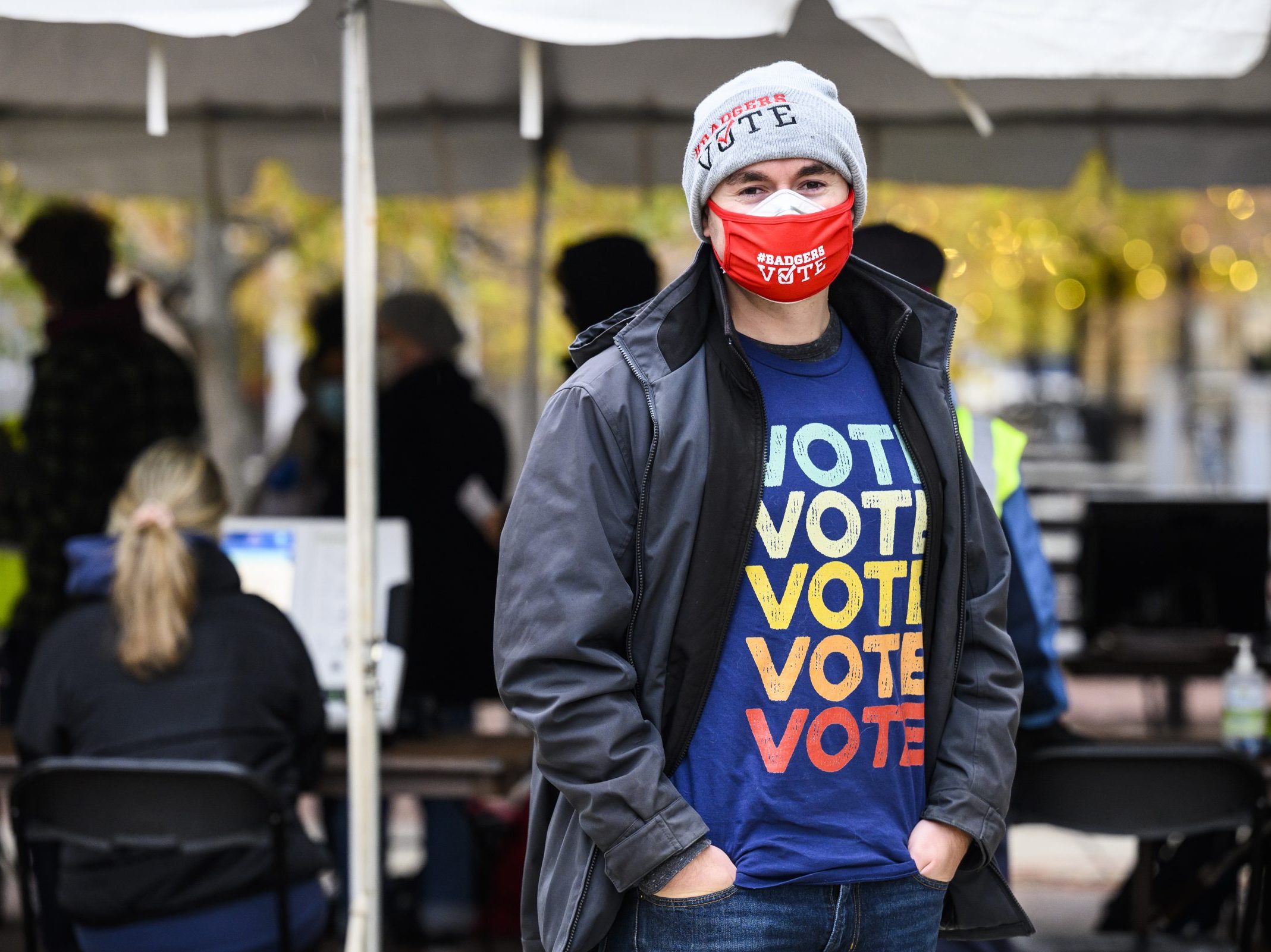 Guy in a mask standing while wearing a shirt with the word "Vote" multiple times stacked on top. Words are different colors going from white on top to red on bottom.